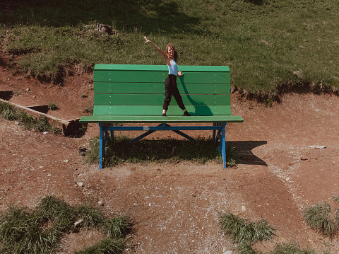 Young Caucasian girl with blonde curly hair performs an athletic yoga pose while standing on a giant green bench surrounded by nature in a mountain setting. The model is smiling and happy