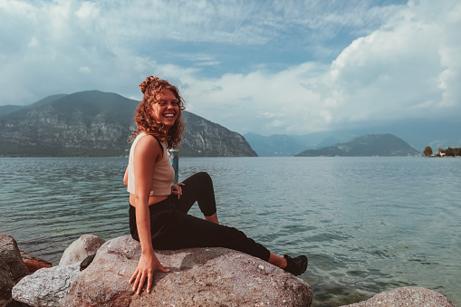 A young girl with light curly hair and summer clothing, smiling pose sitting on the rocks at the water's edge of a lake during summer vacation