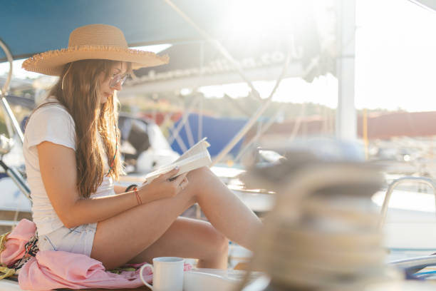 Man with coffee cup is reading book while he is sitting in deck chair on the beach.