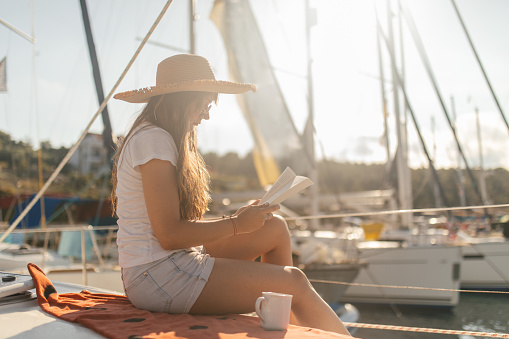 A young woman reading book and drinking coffee on a sailboat. She is enjoying and looking happy and carefree, on sailboat docked in marina in Fiscardo, Kefalonia