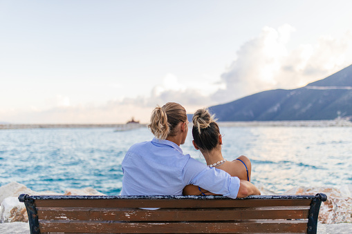 Young couple in love, sitting and hugging on a bench by the sea in a small town in Greece, Vasiliki, Lefkada. They are enjoying while looking at sea before dusk. It's summer, they look carefree and happy.