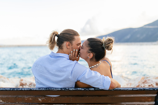 Young couple in love, sitting and kissing on a bench by the sea in a small town in Greece, Vasiliki, Lefkada. It's summer, they look carefree and happy.