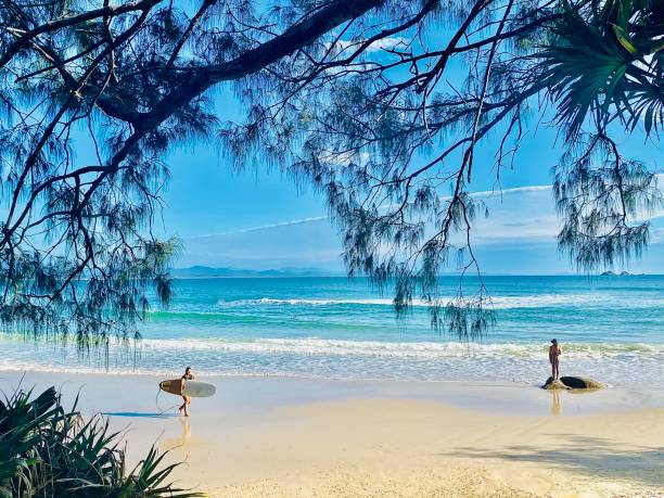 Horizontal seascape of surfer with surfboard walking on sand and person standing on rock looking out to breaking waves on a pastel color clear day at Wategos beach NSW Byron Bay Australia