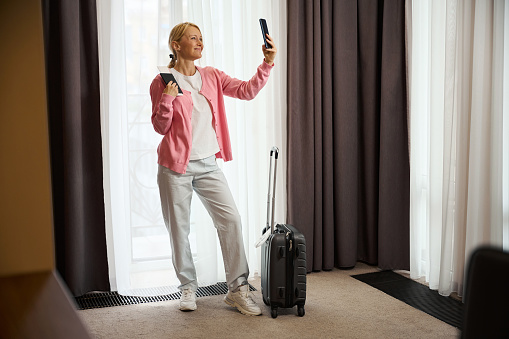 Full-size portrait of joyous female with travel documents in hand posing for smartphone camera against window curtain in suite