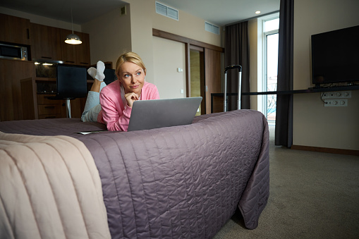 Dreamy lady lying on comfortable bed in suite in front of portable computer