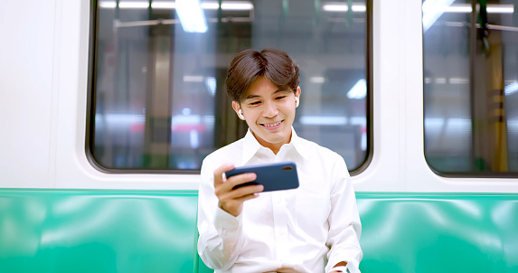 portrait asian businessman is watching videos on live streaming platform through mobile phone and earbuds while sitting commuting at metro mrt car carriage
