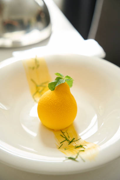 Yellow dessert is served with mint leaves on white plate, cloche lid lies next to it on the serving table