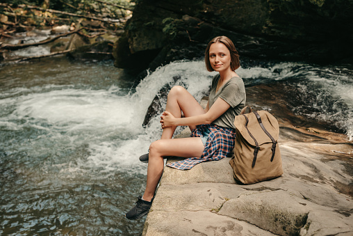 Beautiful female traveler with backpack resting while sitting by the mountain river