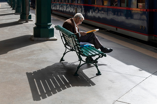 Porto, Portugal-October 3, 2022; Elderly man reading newspaper on a bench on the sunny platform of the São Bento railway station with train at platform; shadow on floor