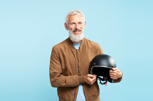Portrait of handsome smiling mature man, gray haired bearded biker holding motorcycle helmet isolated on blue background