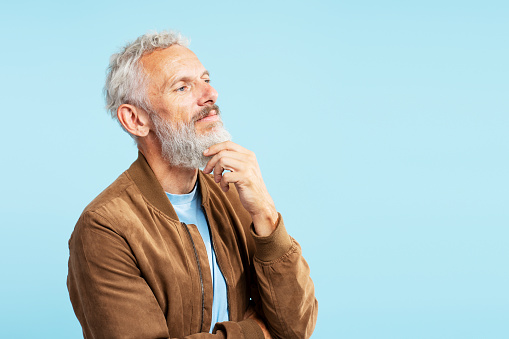 Portrait of pensive serious mature man touching his beard, planning project, brainstorming looking away isolated on blue background