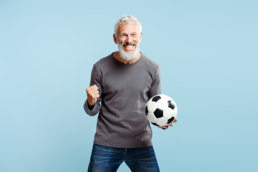 Happy mature man, supporter holding soccer ball celebration success isolated on blue background. Overjoyed male watching football game