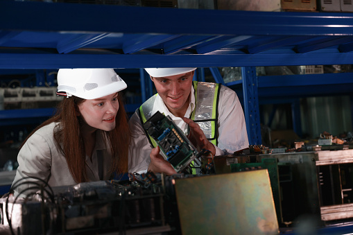 Concentrated male and female engineers in safety gear closely examine electronic components, collaborating on a technical solution in an industrial setting