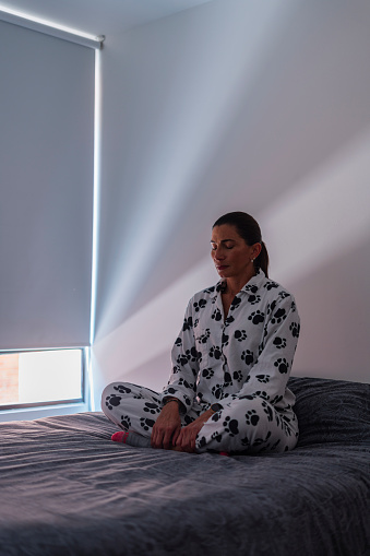 Quiet moment of a Latina woman from Bogota Colombia between 50 and 54 years old, sitting on her bed meditating at the beginning of the day wearing her pajamas