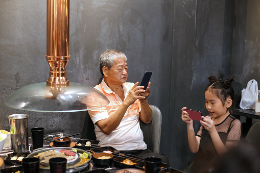Asian family (granddaughter and grandfather) using smartphones while dining in restaurant for barbeque