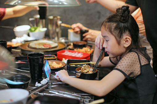 Asian girl distracted with online movie while dining with family in restaurant for barbeque