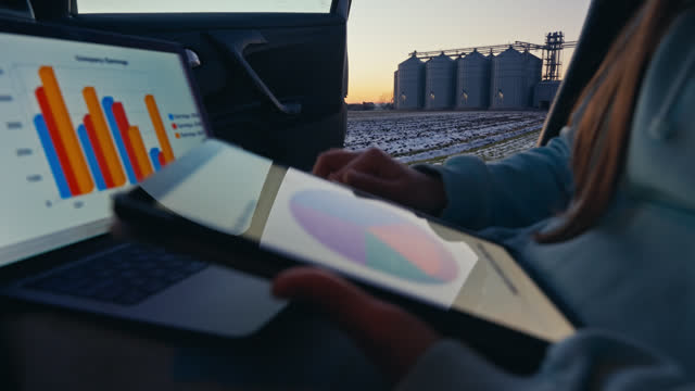 Closeup of Young Female Agronomist Student Analyzing Charts on Tablet and Laptop in Car with Silos in Background During Winter