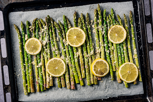 Lemon roasted asparagus fresh out of oven. Simply wash and trim your asparagus stalks, toss them with some olive oil, minced garlic, salt, pepper, parmesan cheese and lemon juice and place the stalks on a piece of baking parchment on an oven tray, add some slices of fresh lemon over the stalks and  bake for around 10 mins at 180 degrees C and, voila delicious and healthy vegetable side dish. Colour, horizontal format overhead view.