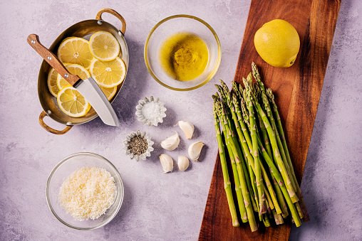 Flat lay of the ingredients needed to make Lemon roasted asparagus fresh out of oven. Simply wash and trim your asparagus stalks, toss them with some olive oil, minced garlic, salt, pepper, parmesan cheese and lemon juice and place the stalks on a piece of baking parchment on an oven tray, add some slices of fresh lemon over the stalks and  bake for around 10 mins at 180 degrees C and, voila delicious and healthy vegetable side dish. Colour, horizontal format overhead view.