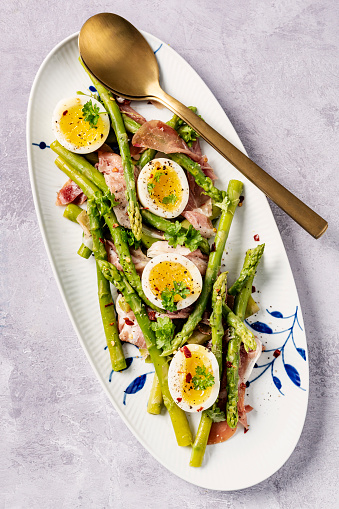 Fresh asparagus paired with jammy eggs and salty prosciutto, ideal for a brunch, lunch or a dinner. Lightly blanch your washed and trimmed asparagus make your vinaigrette with apple cider vinegar, dijon mustard, maple syrup, olive oil, and a shallot. Soft boil your eggs and tear up some prosciutto and combine everything together on a plate. Colour, vertical format overhead view.