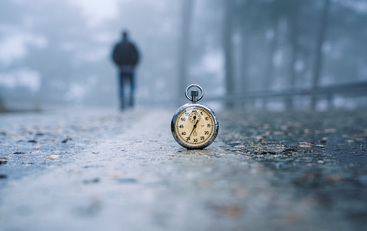 Rear view of a man walks alone on foggy mysterious road, an old stopwatch stands on the road