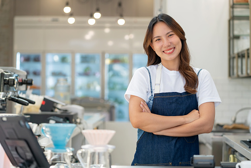 Step into the world of entrepreneurship. Featuring a young coffee shop owner standing confidently in front of the counter, warmly welcoming customers with a genuine smile and arms crossed. Exudes positivity and expertise in her modern coffee shop.
