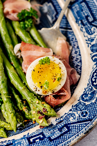 Fresh asparagus paired with jammy eggs and salty prosciutto, ideal for a brunch, lunch or a dinner. Lightly blanch your washed and trimmed asparagus make your vinaigrette with apple cider vinegar, dijon mustard, maple syrup, olive oil, and a shallot. Soft boil your eggs and tear up some prosciutto and combine everything together on a plate. Colour, vertical format overhead view.