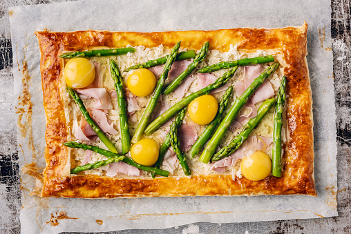 Asparagus tart made with baked eggs and ham. Use shop bought pastry, roll it out and lightly prick it with a fork, apart from the the last 2cm or so from the edge. Lightly bake this for few minutes then add a mix of cream and gruyere cheese to the pastry base, inside the border, add you washed, trimmed and blanched asparagus add the ham and break the eggs in between the asparagus stalks, bake for another 10-15 minutes at 180 degrees C and serve. Colour, horizontal format with a high angle view.
