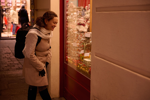 Young multi ethnic woman in warm winter clothes, walking in the city and looking at decorated Christmas showcases at night. Concept of Xmas holidays and preparations. People. Traditions and lifestyles