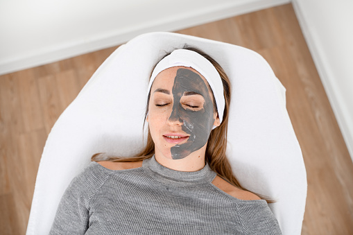 Young female client of a cosmetology salon is lying on a cosmetology couch with a half-face peeling clay mask