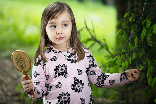 A little girl smiles, looking mysteriously at something off camera, as she walks through a public park in Turin. It is spring, tender green leaves are sprouting on the trees. She holds a small mirror in one hand and a green pine branch in the other, adding a little mystery to the magic of the contest.