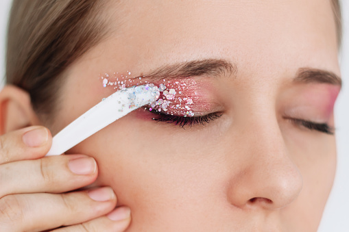 Cropped shot of a young caucasian woman with her eyes closed applying purple and pink eye shadow and glitter on the eyelids. Festive party, holiday makeup. Close up
