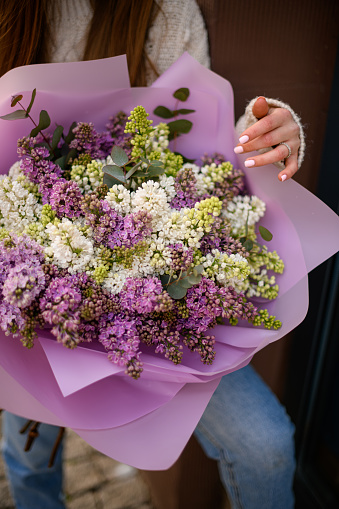 Selective focus on huge bouquet of colorful lilac flowers in paper package in woman's holding in hands. Street photo. Florist shop. Concept of bloom, holiday, gift