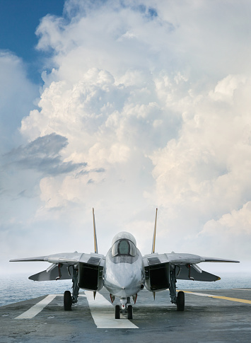 An F-14 jet fighter on an aircraft carrier deck beneath dramatic clouds viewed from front