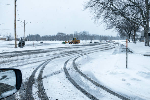 Tire tracks and a distant front loader snowplow parked in a snowy winter blizzard suburban parking lot.