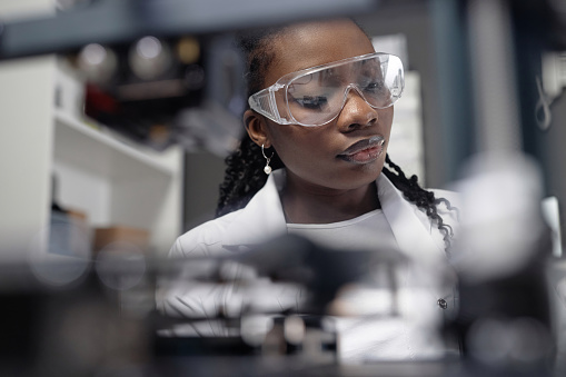 Look through blurred 3D printer components of young Black woman laboratory specialist in protective goggles