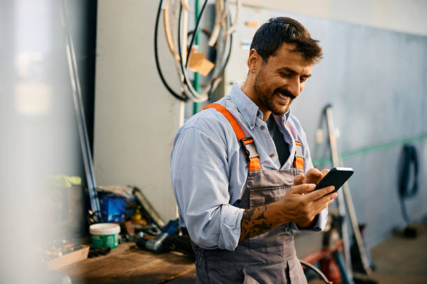 Happy mechanic using smart phone while working in a repair shop. Happy mechanic texting on cell phone while working in a garage. Copy space. small business stock pictures, royalty-free photos & images