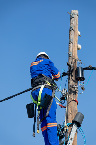 electrician wearing a belt and hardhat on a ladder on a wooden pole repairing electricity
