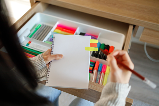 A person selects a marker from a drawer full of colorful stationery, holding a notepad with sticky notes.