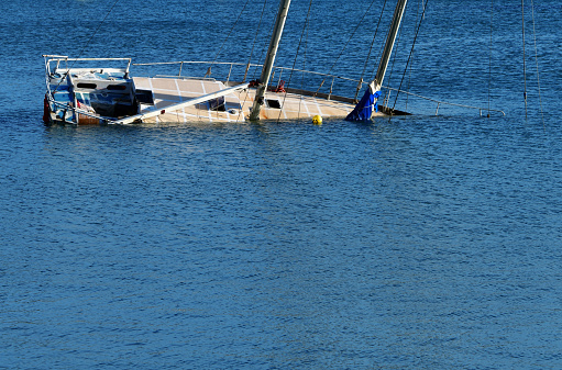 A two mast sailboat lies capsized in tranquil blue waters, with its mast and sails partially submerged, indicating a recent incident or abandonment. Starboard view.