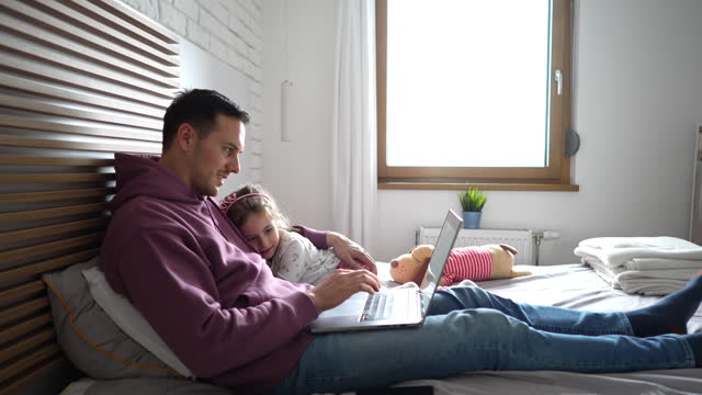 Young father working on laptop, while lying in the bed with his daughter