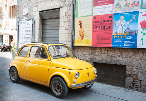 Gubbio Italy - May 13 2011; old yellow small Italian vintage car parked in old town street under wall of posters in Italian language.