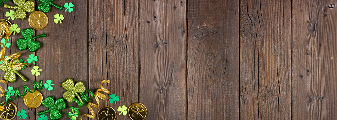 St Patricks Day corner border of green shamrocks, gold coins and ribbon. Top down view over a rustic dark wood banner background with copy space.