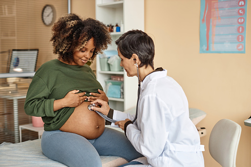 Smiling young African American expectant mother sitting on medical table in clinic while female obstetrician using stethoscope listening baby