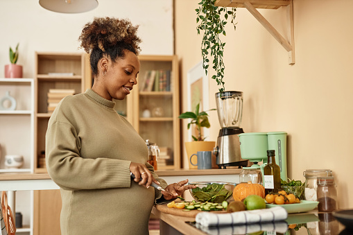 Side view of young pregnant Black woman with curly bun making healthy dinner with vegetables at home