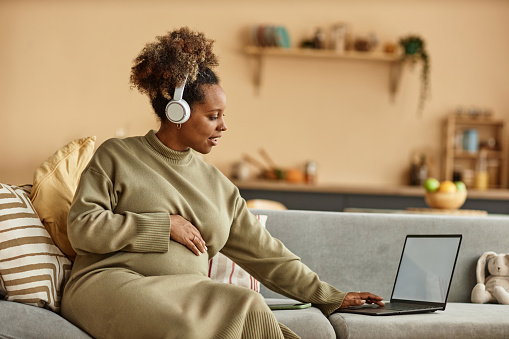 Medium long shot of young Black expectant mother in headphones sitting on couch at home while working on laptop with blank screen
