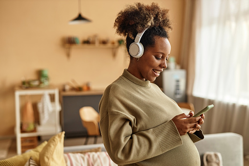 Medium shot of smiling pregnant Black woman at home wearing headphones holding smartphone and texting with smartphone