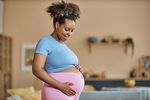 Medium shot of happy pregnant Black woman at home holding her round belly standing in blue crop top and pink trousers while looking down