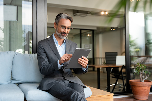 Smiling mature European Latin businessman holding tablet gadget sitting in office with copy space. Middle aged man manager using digital device app. Technology application and solutions for business.