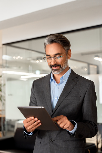Vertical portrait of smiling Hispanic mature adult professional business man, happy Indian 40s 50s businessman CEO holding digital tablet using fintech tab application standing inside company office.